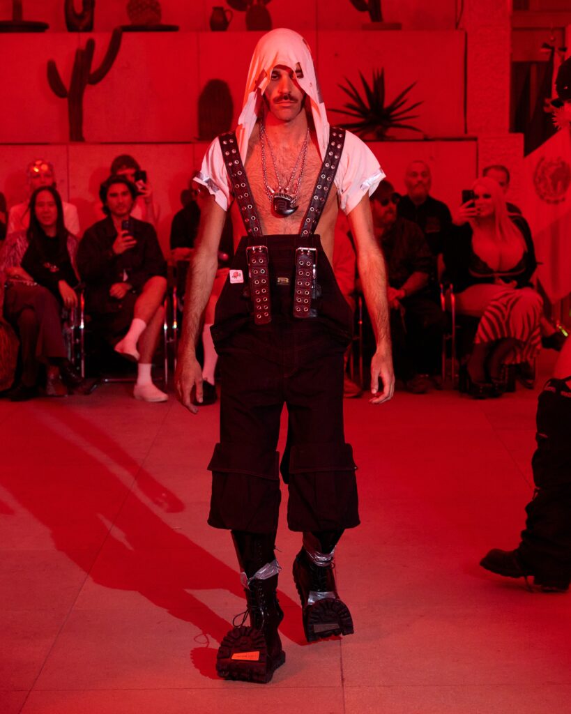 Male model walks a runway in a white hooded shirt with studded leather harness, chains, and chunky boots under red lighting, audience in background