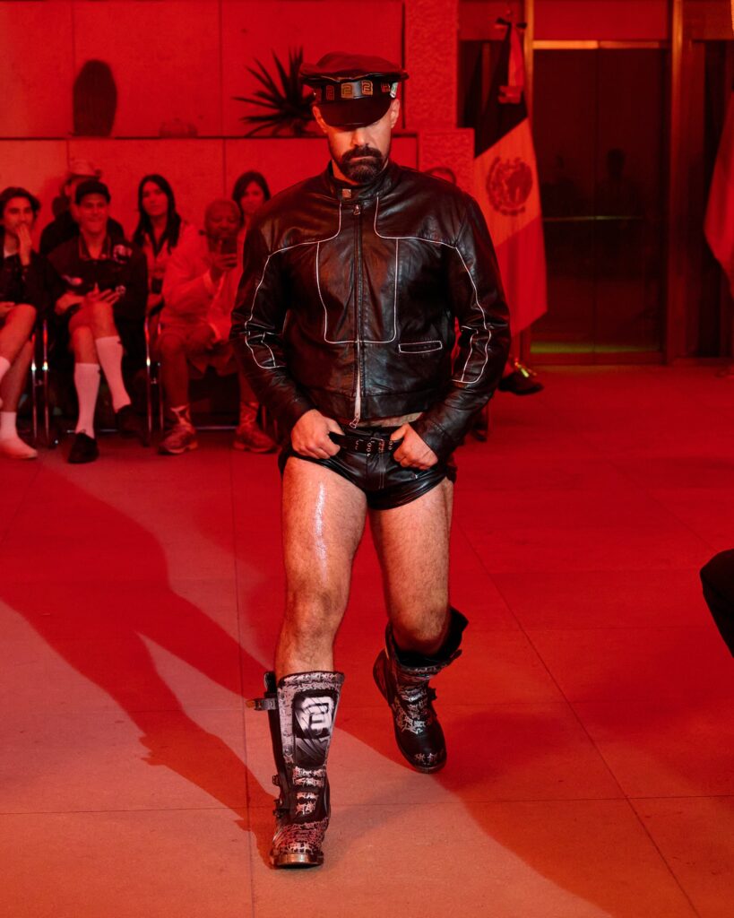 Male model in black leather outfit and cap walks the runway; wearing knee and shin armor with roller skates, red lighting and seated audience in background.