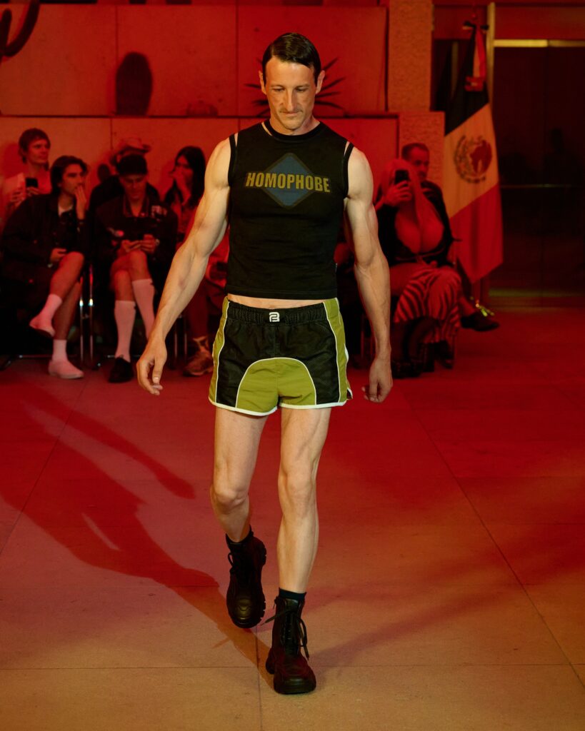 Male model walks a red-lit runway wearing a black sleeveless shirt that reads HOMOPHOBE, with spectators in the background watching.