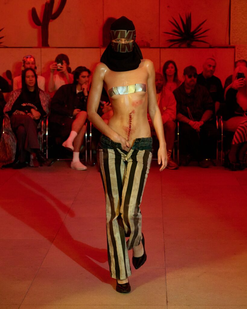 Model walks a fashion show wearing a black head covering with a metallic face wrap and vertically striped pants, under red lighting with seated spectators in the background.