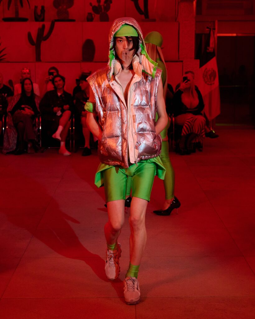 Model walking a fashion show in a shiny silver puffer vest with a green hood, lime-green shorts, and red stage lighting; audience seated in the background.