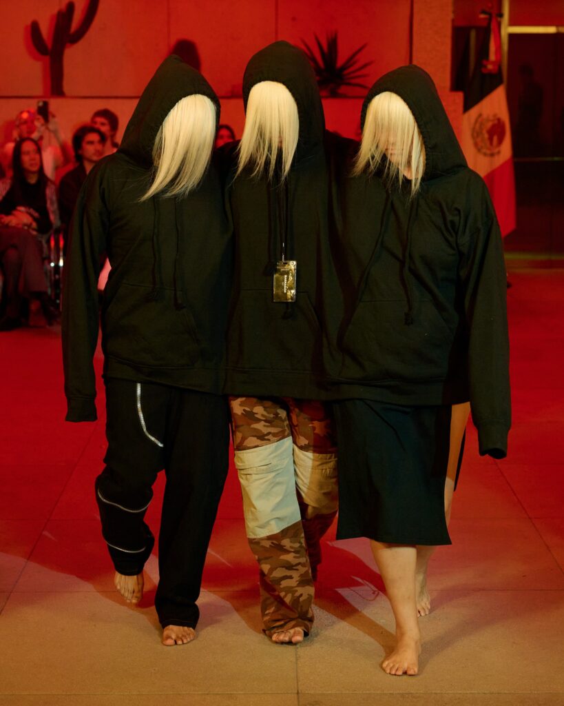 Three people in black hooded robes with long blond wigs stand close together on a red-lit stage.