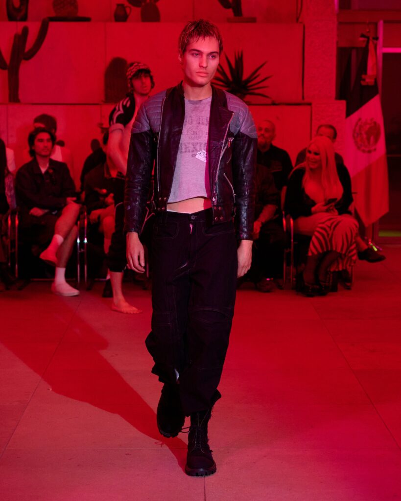 Male fashion model walks down a runway wearing a black leather jacket and dark pants, red lighting and an audience in the background.