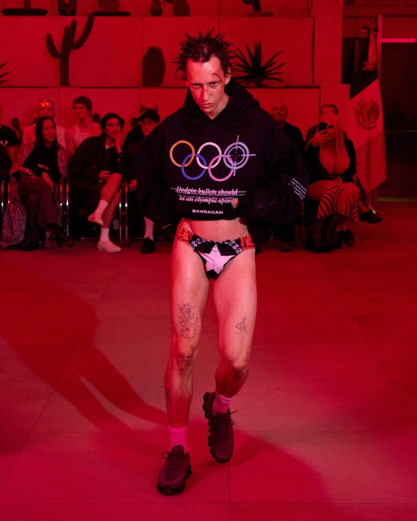 Male model walks a fashion runway in a black hoodie with a colorful Olympic rings graphic, red lighting, crowds watching in the background, tattoos on legs.