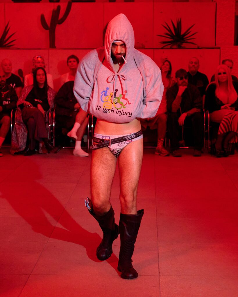 Wrestler in a gray hooded sweatshirt and brief trunks, cigarette in mouth, standing on a red-lit stage with seated audience behind him.