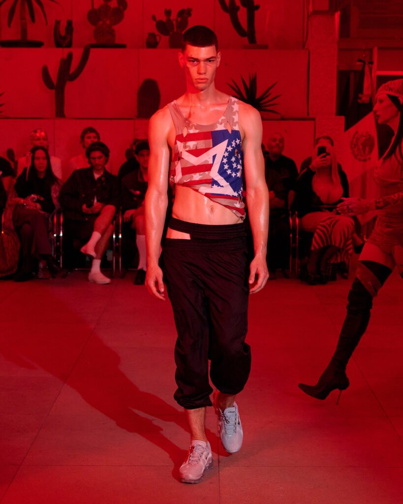 Male model walks a runway in a sleeveless American flag print top and black pants, under red lighting with audience in the background.