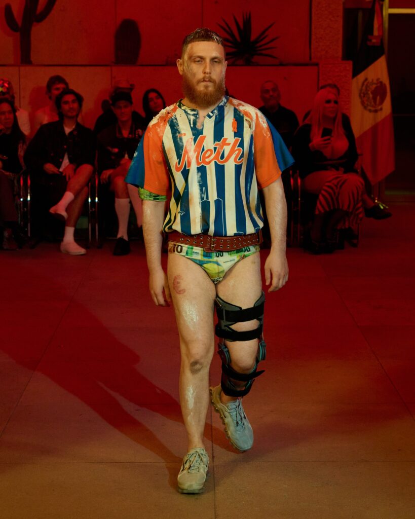 Bearded man walks a runway in a blue‑white striped shirt with 'Meth' logo, knee braces, red lighting, and seated audience in the background.