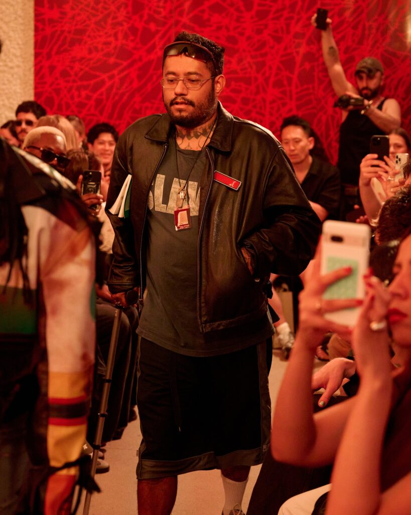 Man in a black leather jacket walks through a crowded event, holding papers; red abstract wall in background, spectators with cameras.