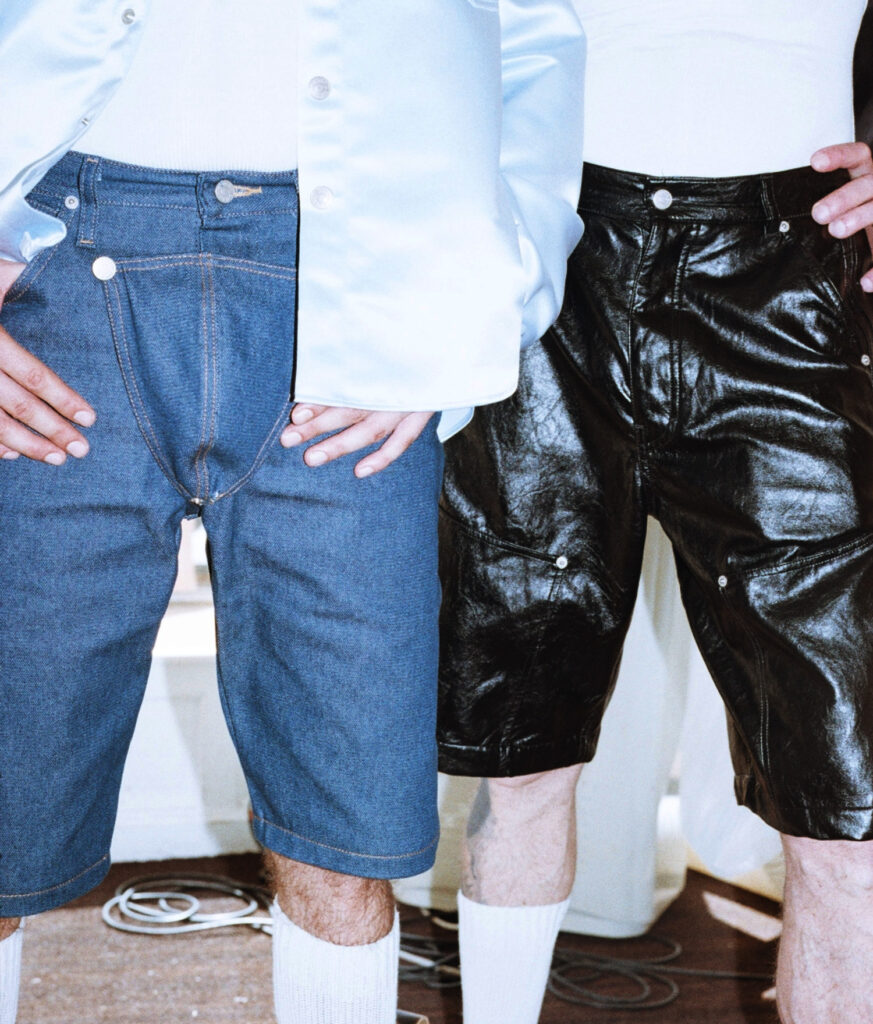 Two people standing side by side wearing casual outfits: blue denim shorts and a white shirt on the left, shiny black pants on the right, with hands on hips.
