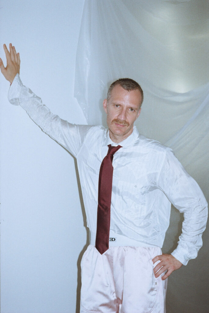 Man in a white dress shirt and red tie with one arm raised, posing against a white backdrop.