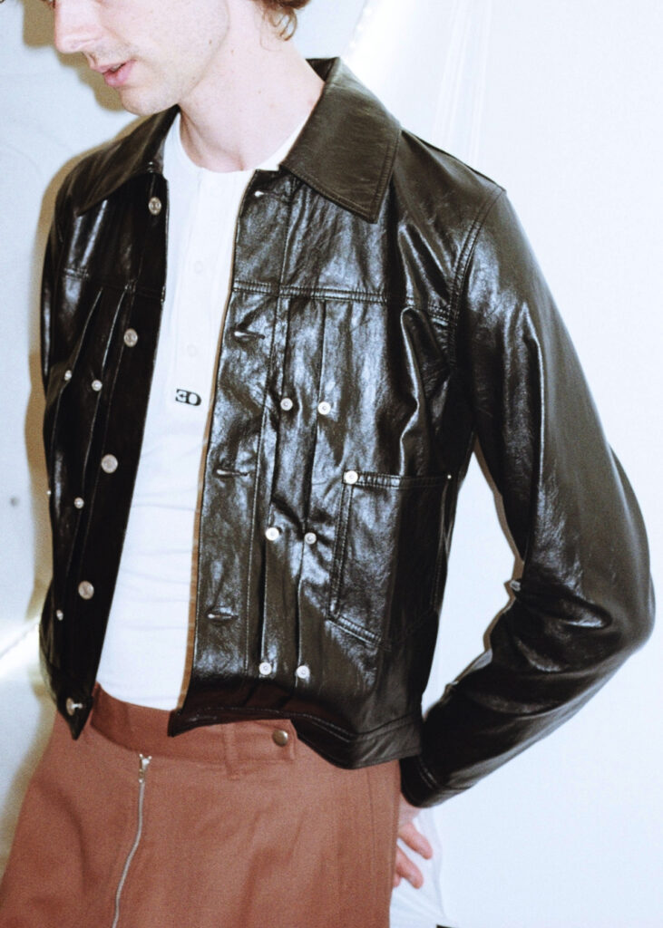 Person wearing a shiny black leather jacket over a white shirt, posing against a light background in a fashion shot.