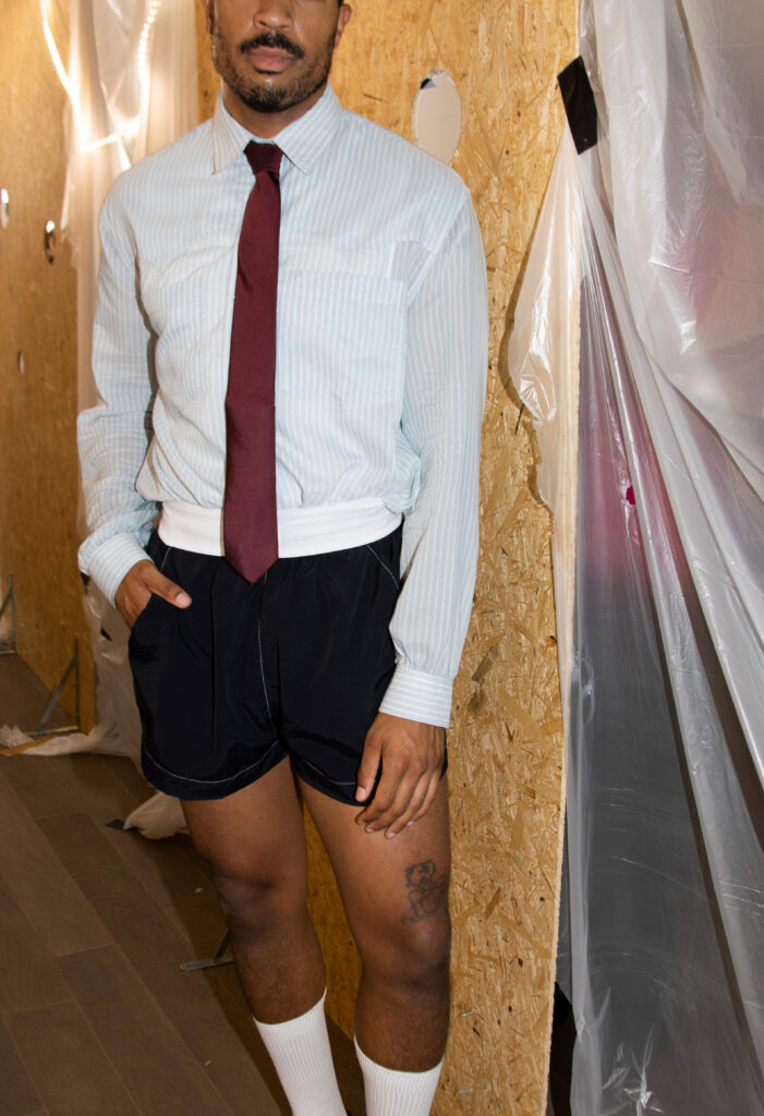 Man in a light blue striped dress shirt and maroon tie standing next to a plywood wall with plastic sheeting in a construction area.