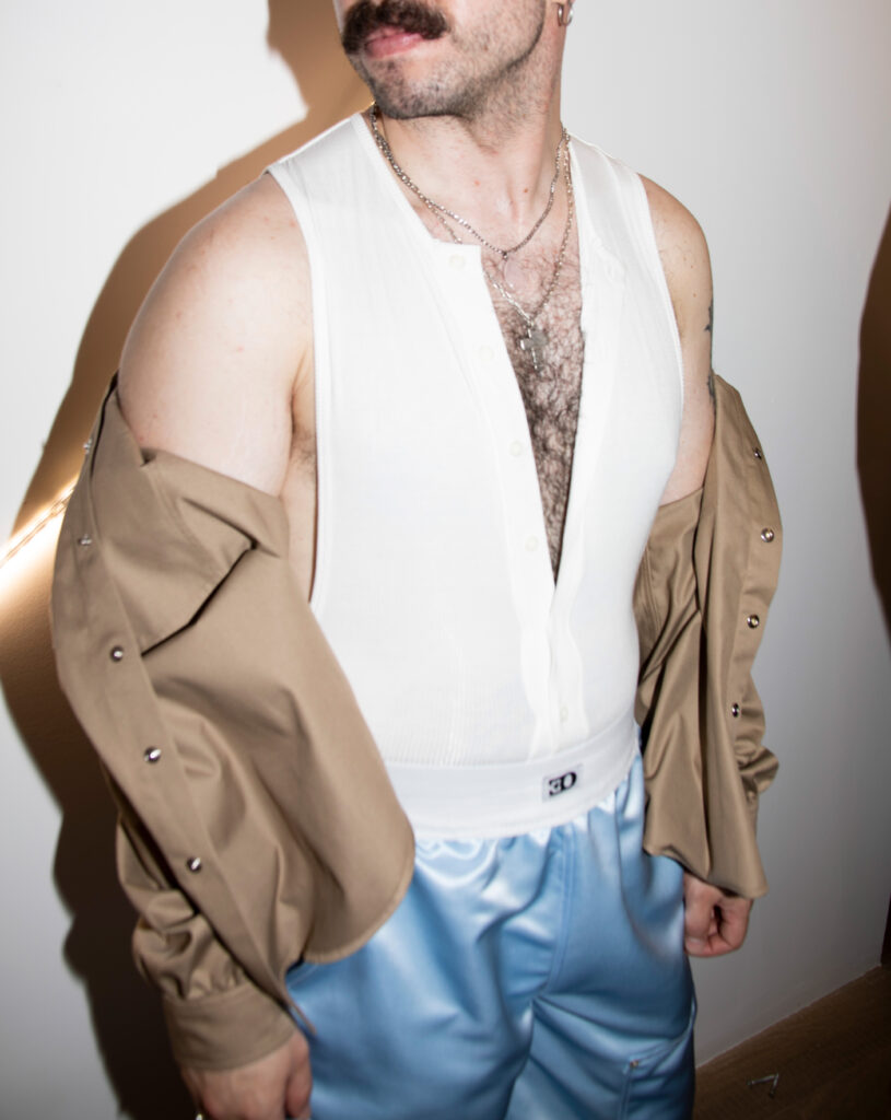 Torso of a person wearing a white sleeveless top, beige jacket draped off the shoulders, layered silver necklaces, and light blue satin pants.