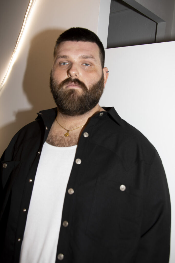 Bearded man wearing a black button-down shirt over a white tee, gold necklace, standing indoors against a white wall and doorway.