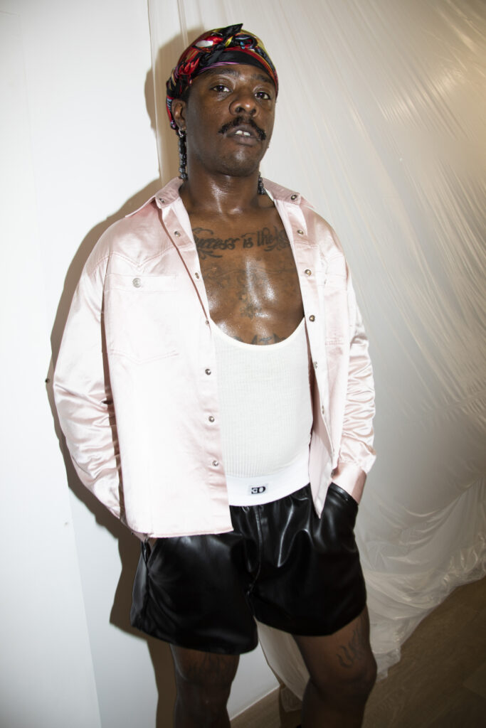 Man with a colorful head wrap, pink unbuttoned shirt over a white tank top, black shorts, and gloves posing against a white plastic backdrop.