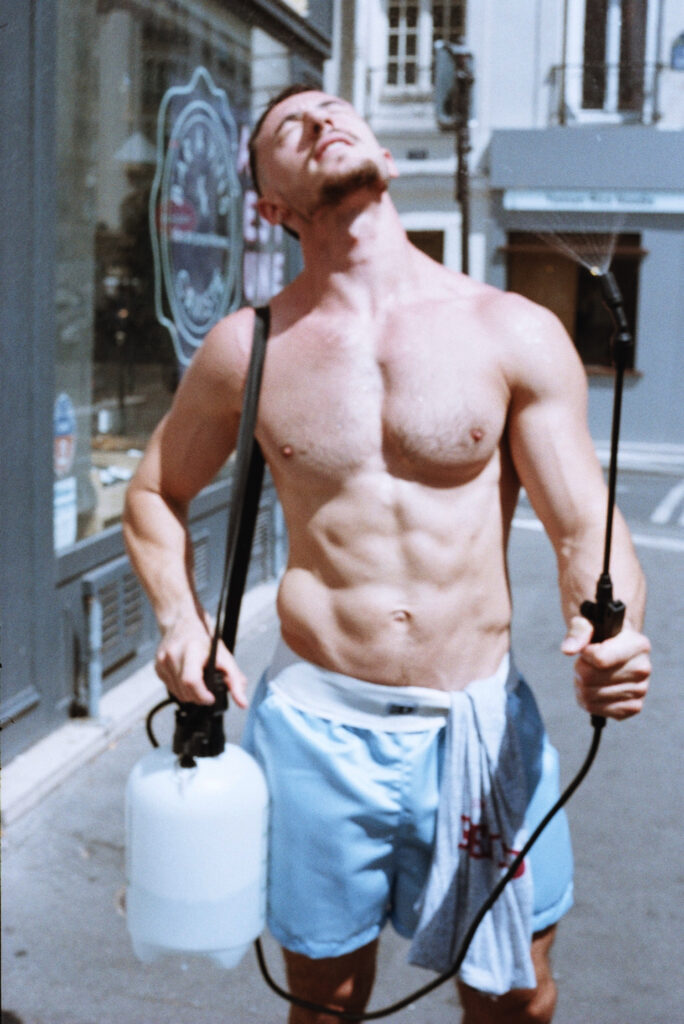 Shirtless man in light blue shorts on a city street, carrying two scuba tanks with hoses and looking upward.