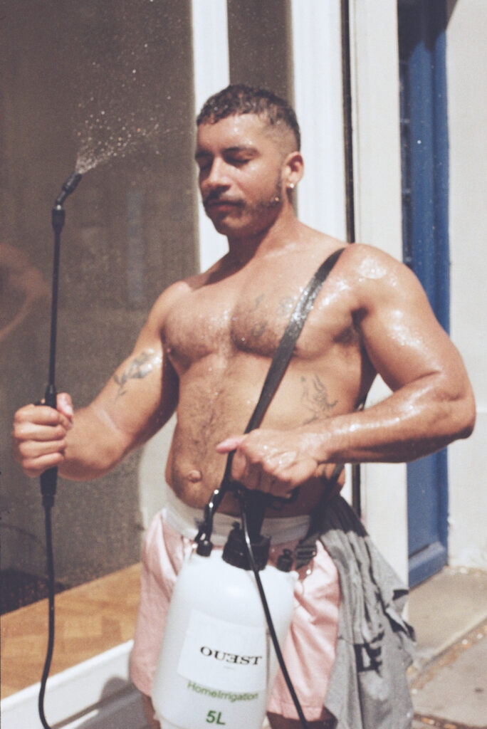 Shirtless man with short hair and earrings, wearing a chest strap, holding a water hose as water sprays outward outdoors near a doorway.