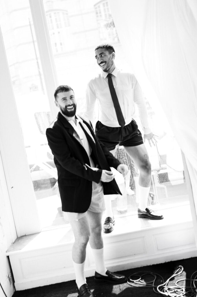 Two men in business attire smiling inside a bright glass-walled room; one wearing a suit jacket, the other in a dress shirt and tie with shorts.