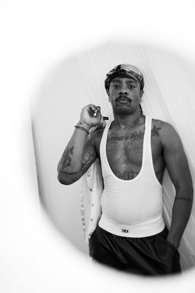 Black-and-white portrait of a tattooed man wearing a bandana, white tank top, and gold bracelets, facing the camera.