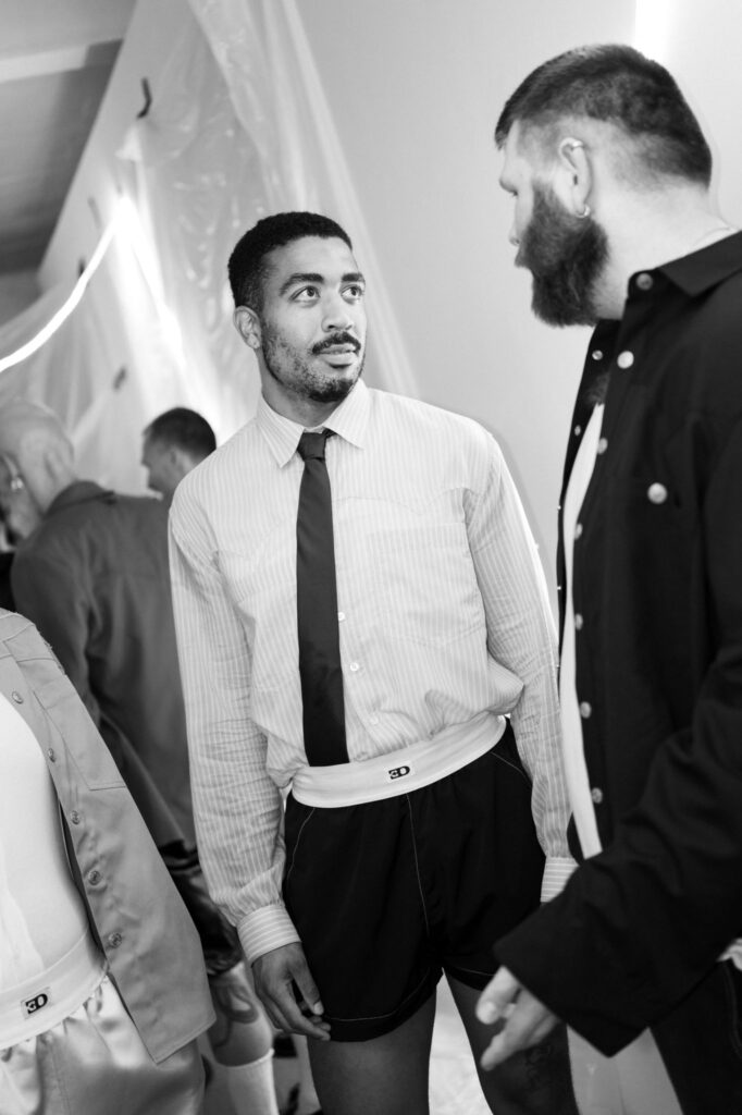 Two men in formal attire converse at an indoor event; one wearing a striped shirt and tie, the other with a dark jacket and beard.