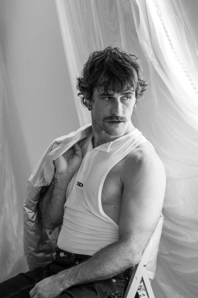 Black-and-white portrait of a man with a mustache and short hair, wearing a white sleeveless shirt, seated beside a draped backdrop, looking to the side.