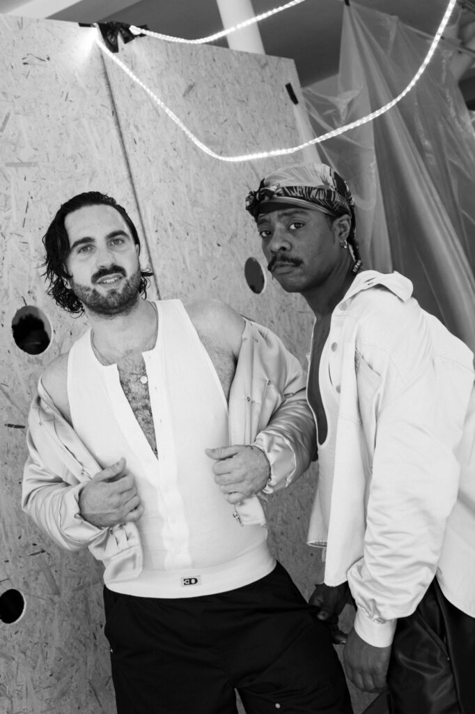 Two men pose together in a white jackets and tank tops against a plywood wall, one with a bandana, looking at the camera.