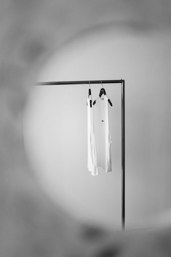 White shirts on hangers hanging from a simple metal clothing rack in a softly blurred room, minimal and stylish. Racks and garments are the focus behind a large blurred foreground element.