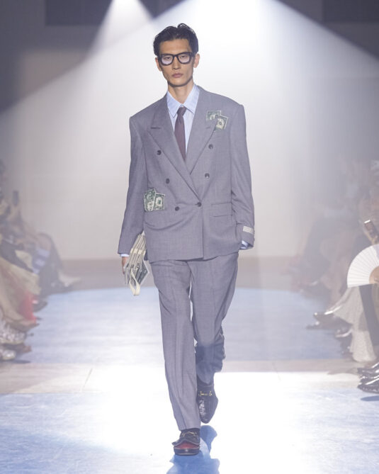 Silhouette doublet SS27 portant un costume gris croisé, une chemise rayée, une cravate et des détails façon billets cousus sur la veste.