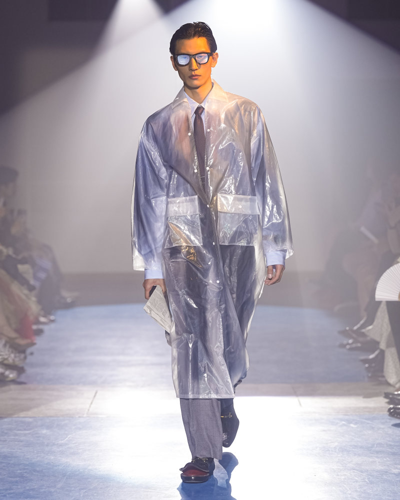 Silhouette doublet SS27 portant un manteau transparent sur une chemise rayée, une cravate, un pantalon gris et des lunettes.